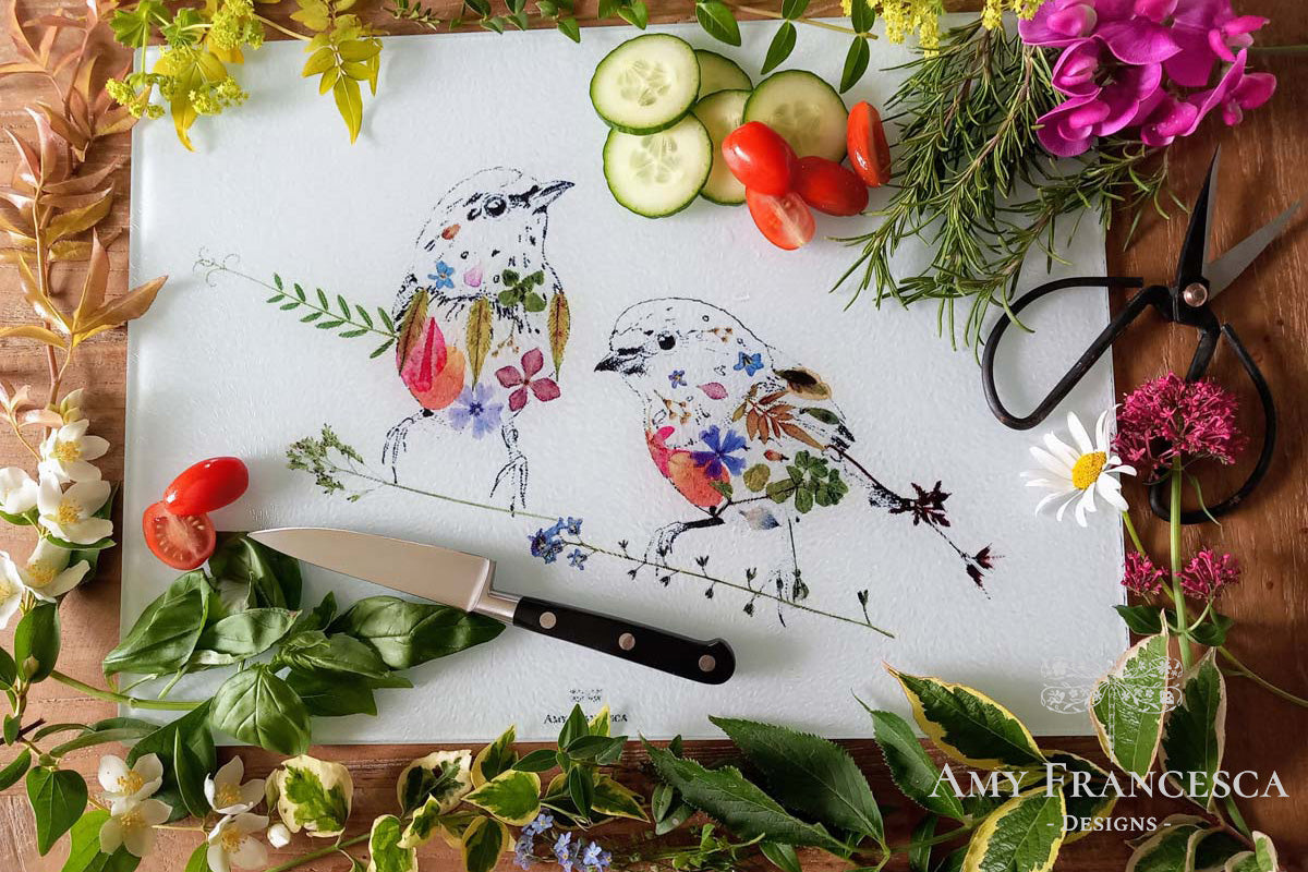 Chopping board featuring two bird illustrations, shown with sliced vegetables, herbs and surrounding flowers.