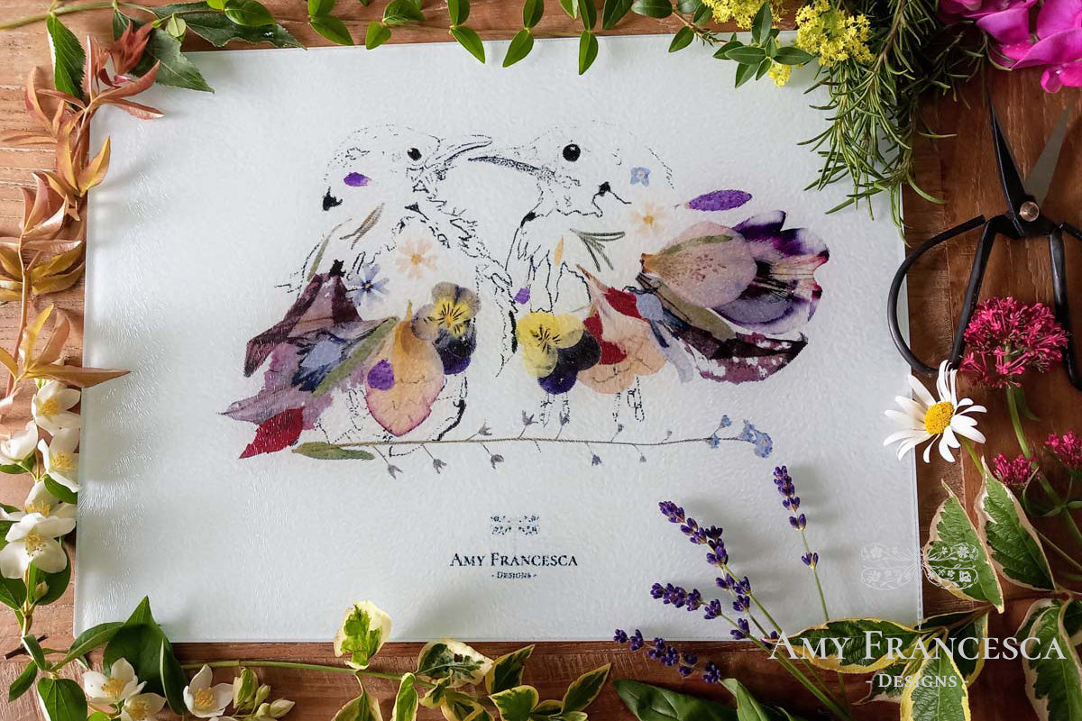Chopping board featuring two bird illustrations, shown with surrounding flowers, foliage and scissors.