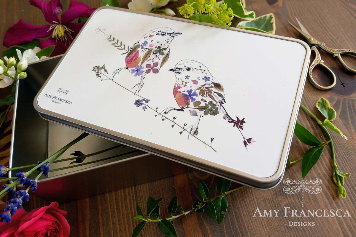 Rectangular storage tin featuring two bird illustrations, shown with surrounding flowers and scissors.