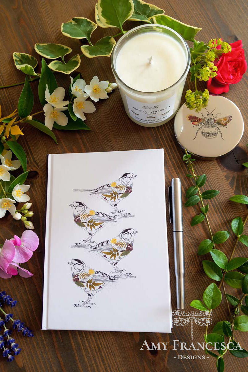 Notebook featuring four bird illustrations on a white cover, shown with a candle, flowers, leaves and a pen.