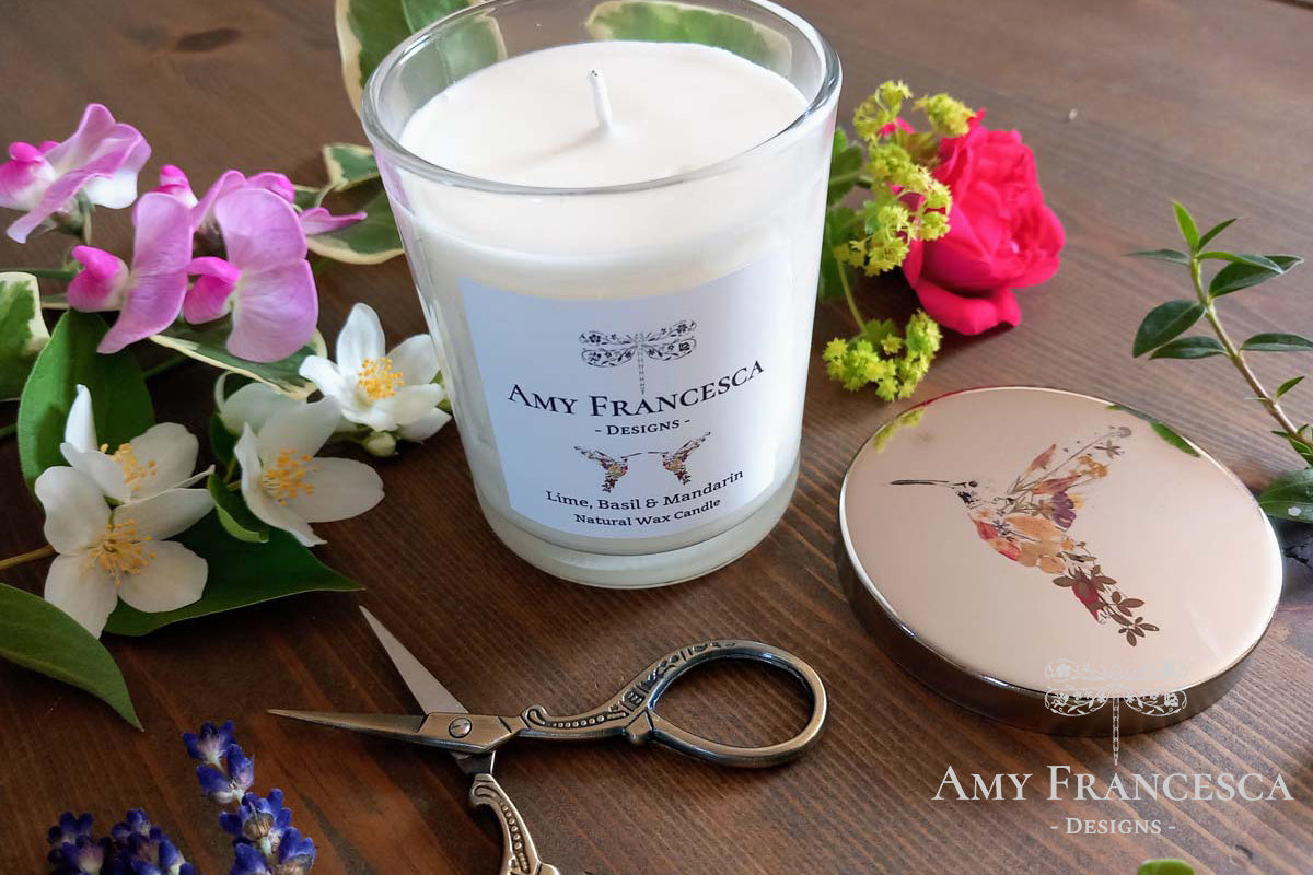 Candle featuring hummingbird illustrations on both the lid and the jar label, shown with surrounding flowers and scissors.