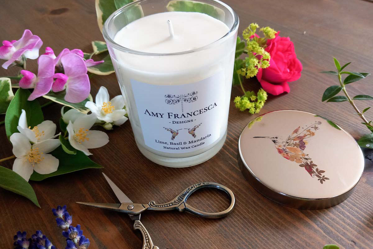 White candle with branded label, shown with a hummingbird‑illustrated lid and assorted flowers.