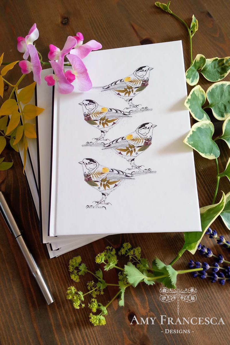 Stacked notebooks with the top one featuring four bird illustrations on a white background, shown with surrounding flowers and a pen.