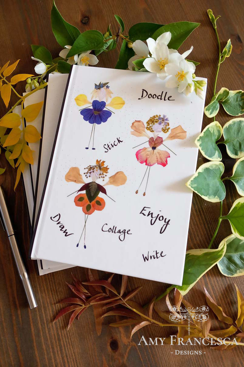 Stacked notebooks with the top one featuring floral figures with wings on a white cover with the words “Doodle,” “Stick,” “Draw,” “Collage,” “Enjoy,” and “Write,” shown with surrounding flowers, leaves and a pen.
