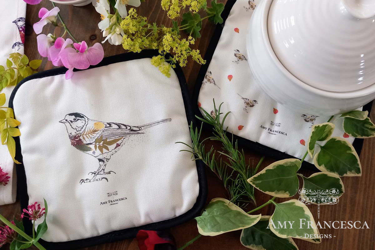 Two pot holders featuring bird illustrations, shown with a ceramic dish and surrounding flowers and greenery.