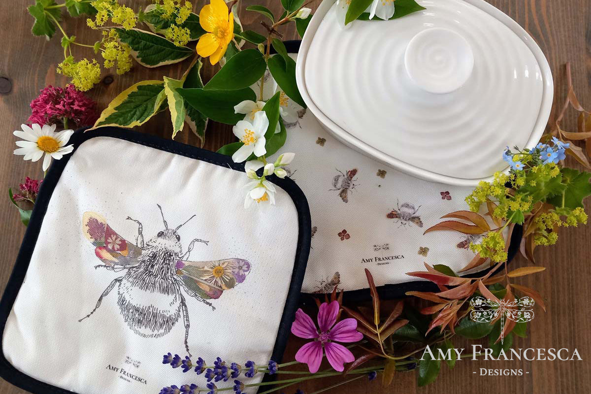 Two pot holders featuring bee illustrations, shown with a ceramic dish and surrounding flowers.