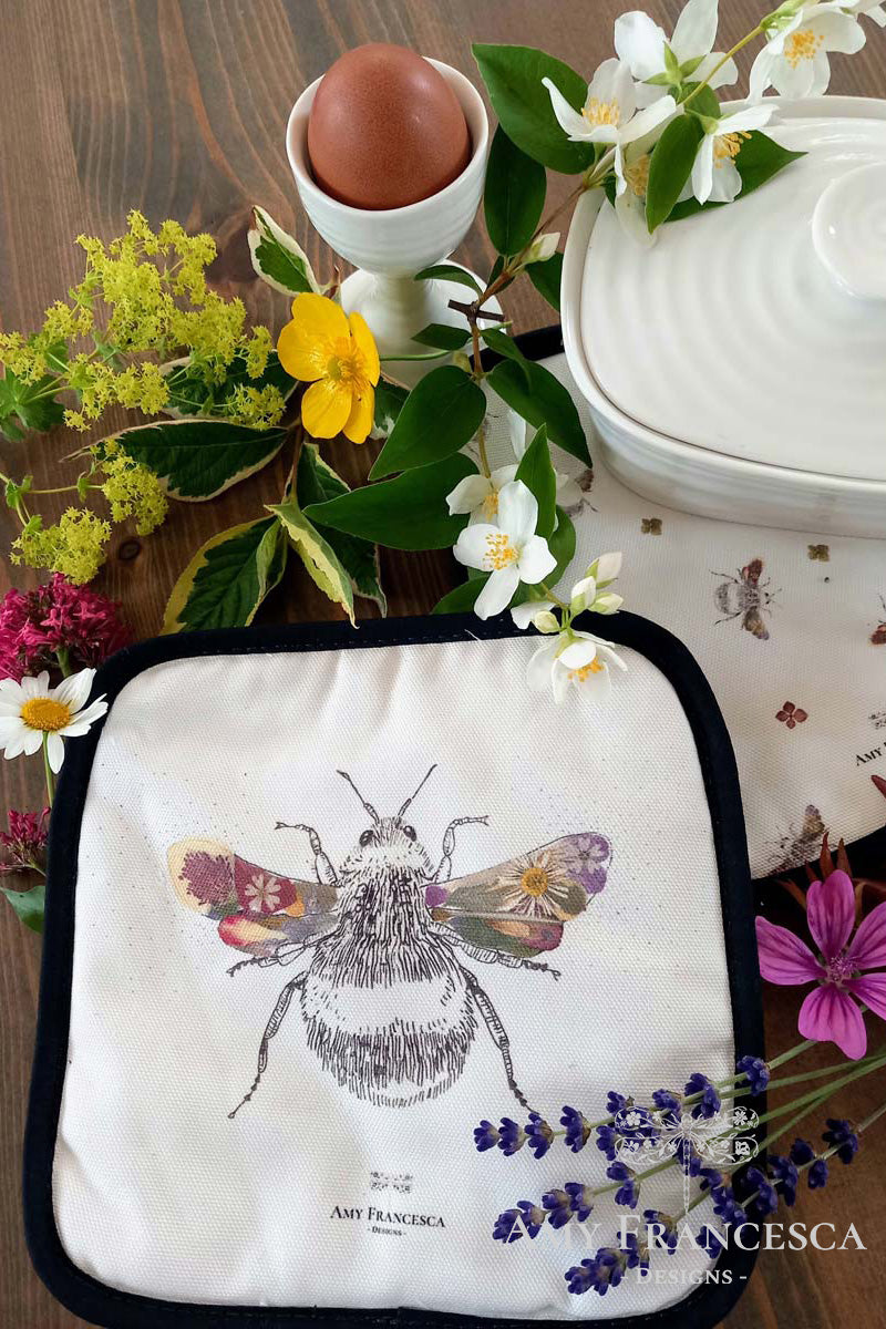 Pot holder pair featuring a bee illustration, shown with an egg cup, dish and assorted flowers.
