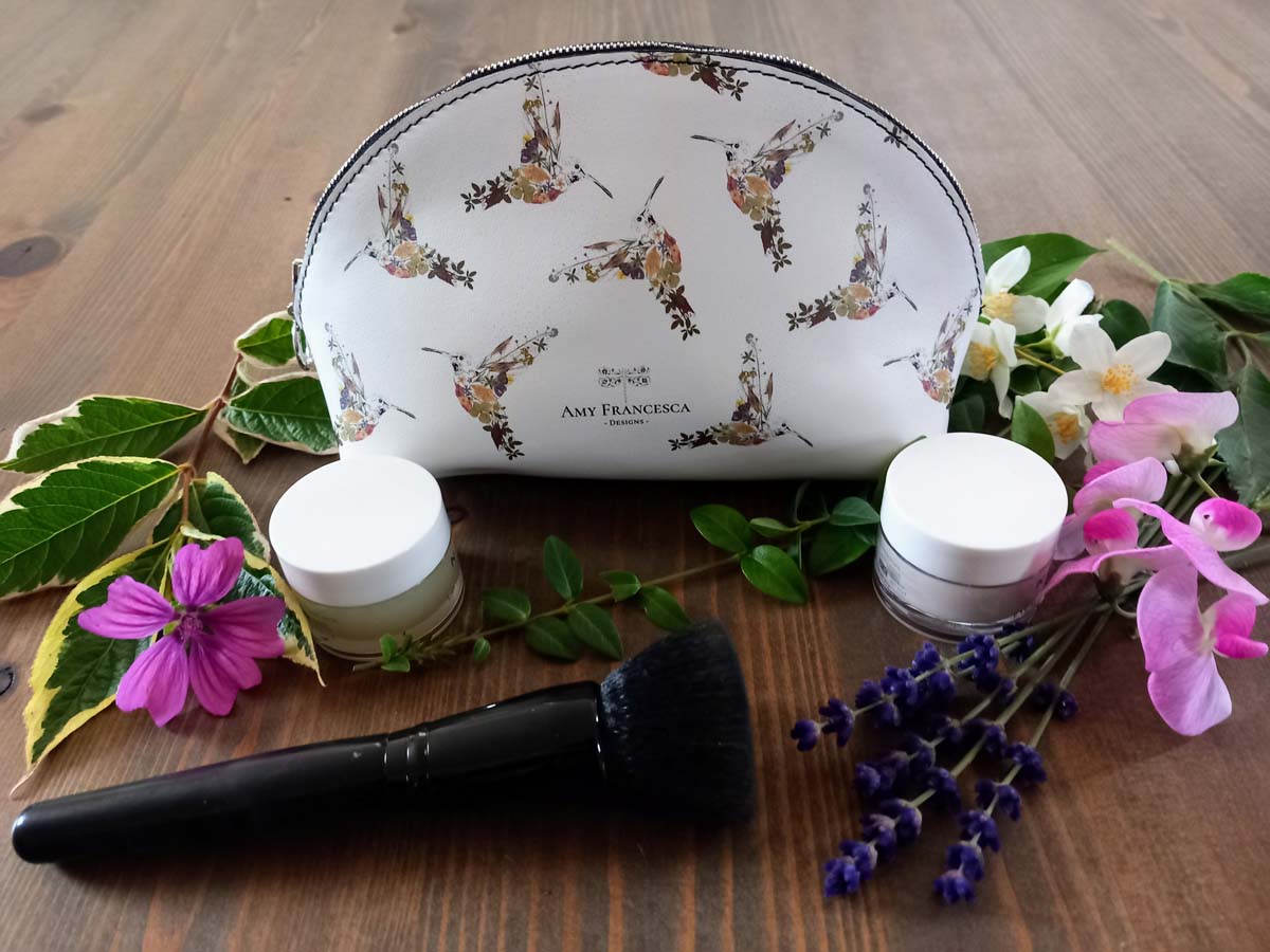 Cosmetic case featuring a repeating hummingbird illustration, shown with flowers, greenery, small jars and a makeup brush.