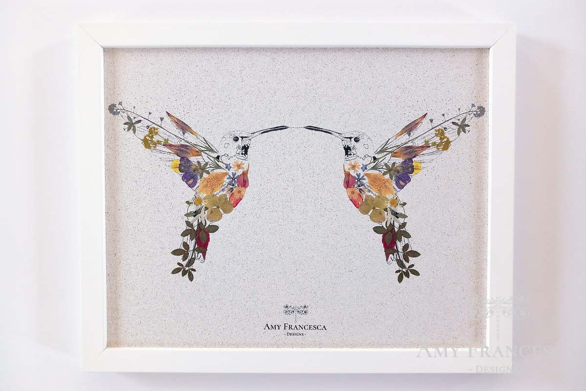 A white framed cork pin board featuring an illustration of two hummingbirds facing each other, placed on a light background.