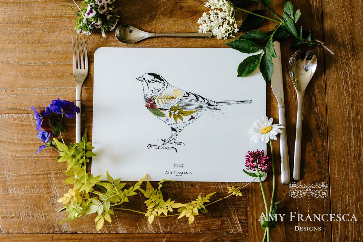 Placemat featuring a bird illustration, shown with cutlery and surrounding flowers.