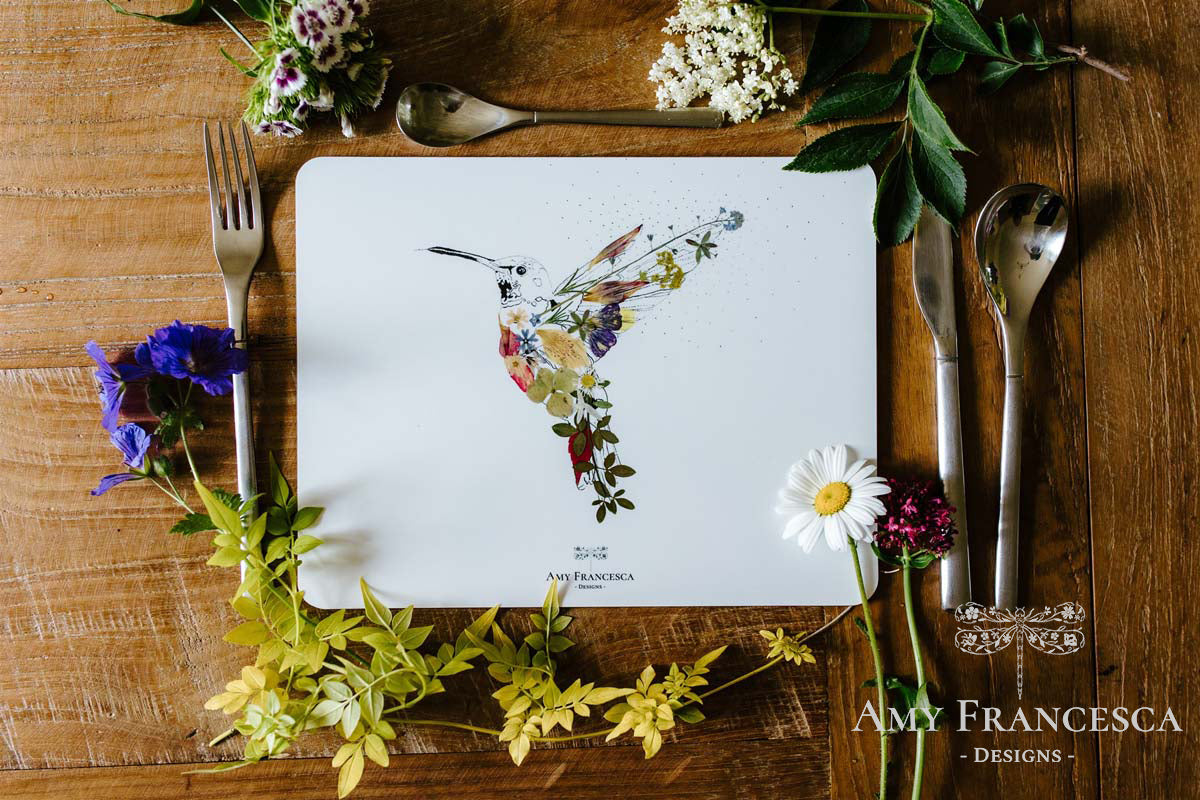 Placemat featuring a hummingbird illustration, shown with cutlery and surrounding flowers.