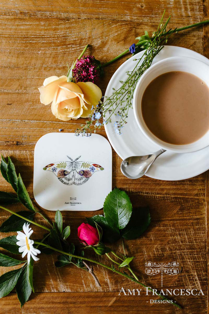 Coaster featuring a moth illustration, shown beside a cup and assorted flowers.