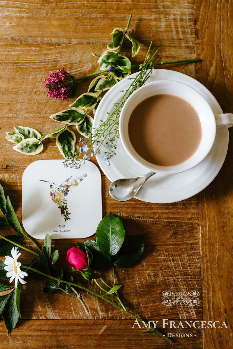 Coaster featuring a hummingbird illustration, shown beside a cup and surrounding flowers.
