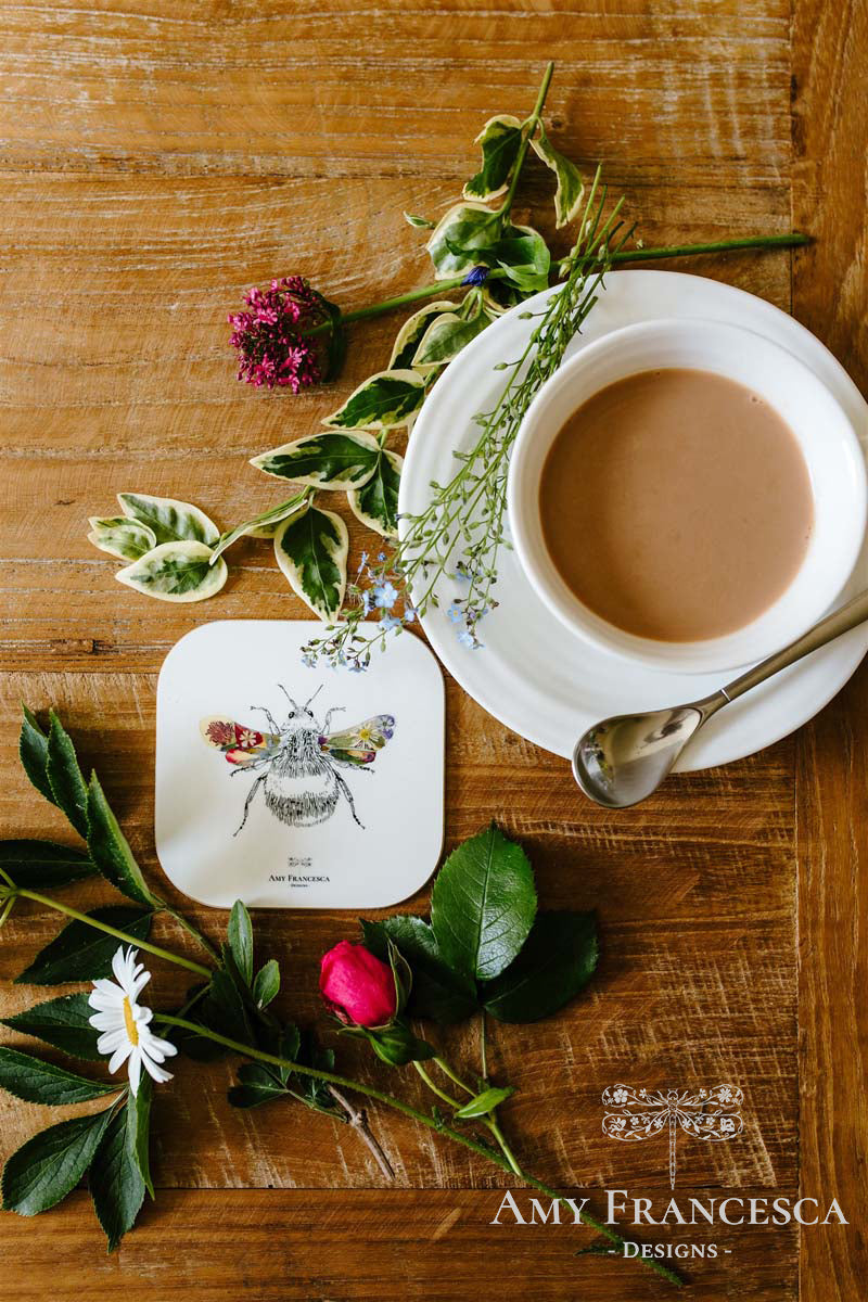 Coaster featuring a bee illustration, shown beside a cup and surrounding flowers.