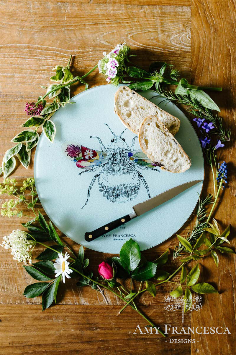 Chopping board featuring a bee illustration, shown with sliced bread, a knife and surrounding flowers.