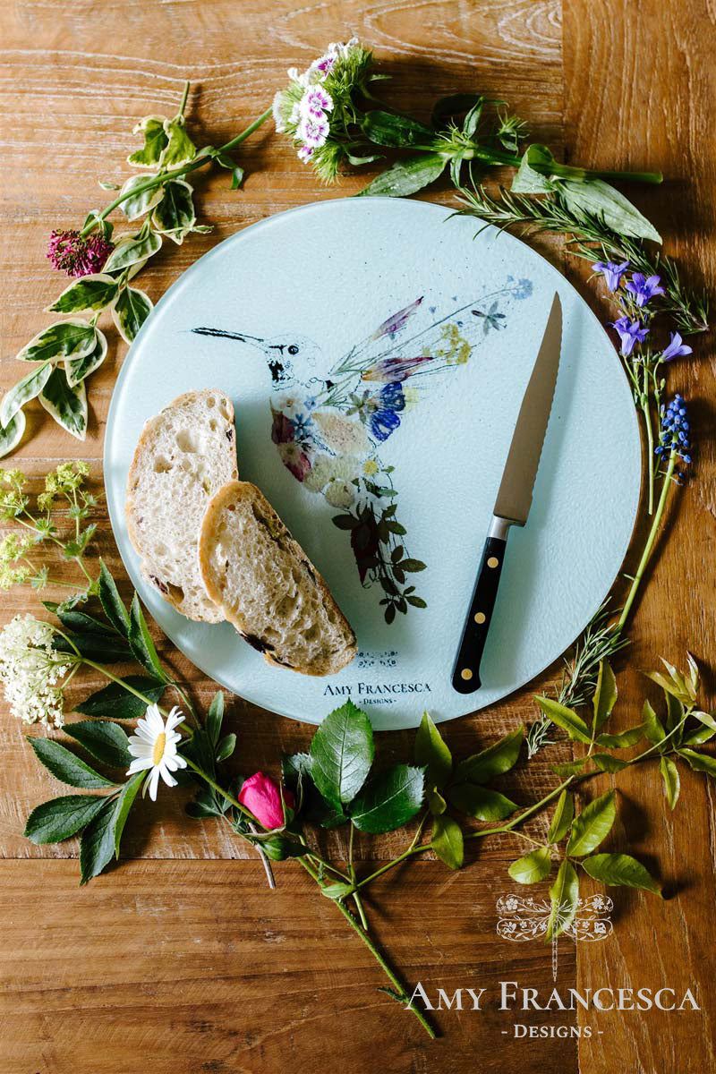 Chopping board featuring a hummingbird illustration, shown with sliced bread, a knife and surrounding flowers.