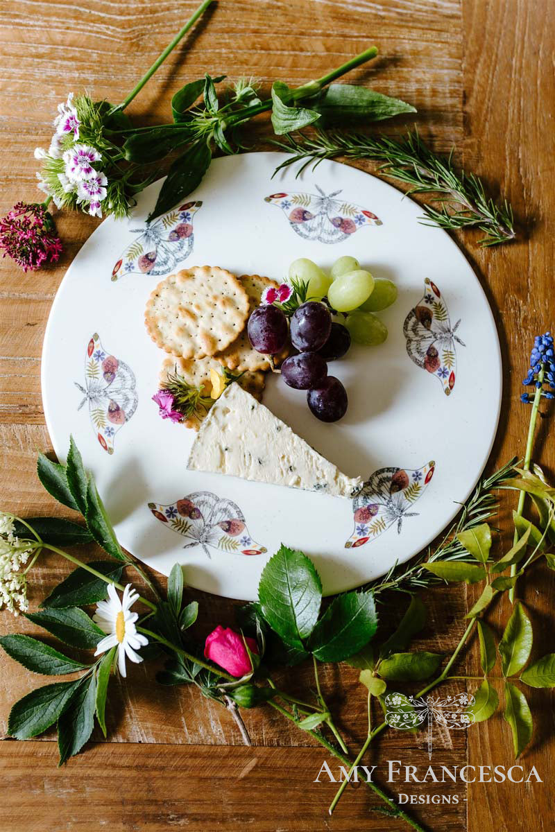 Serving platter featuring moth illustrations, shown with cheese, grapes and crackers surrounded by flowers.