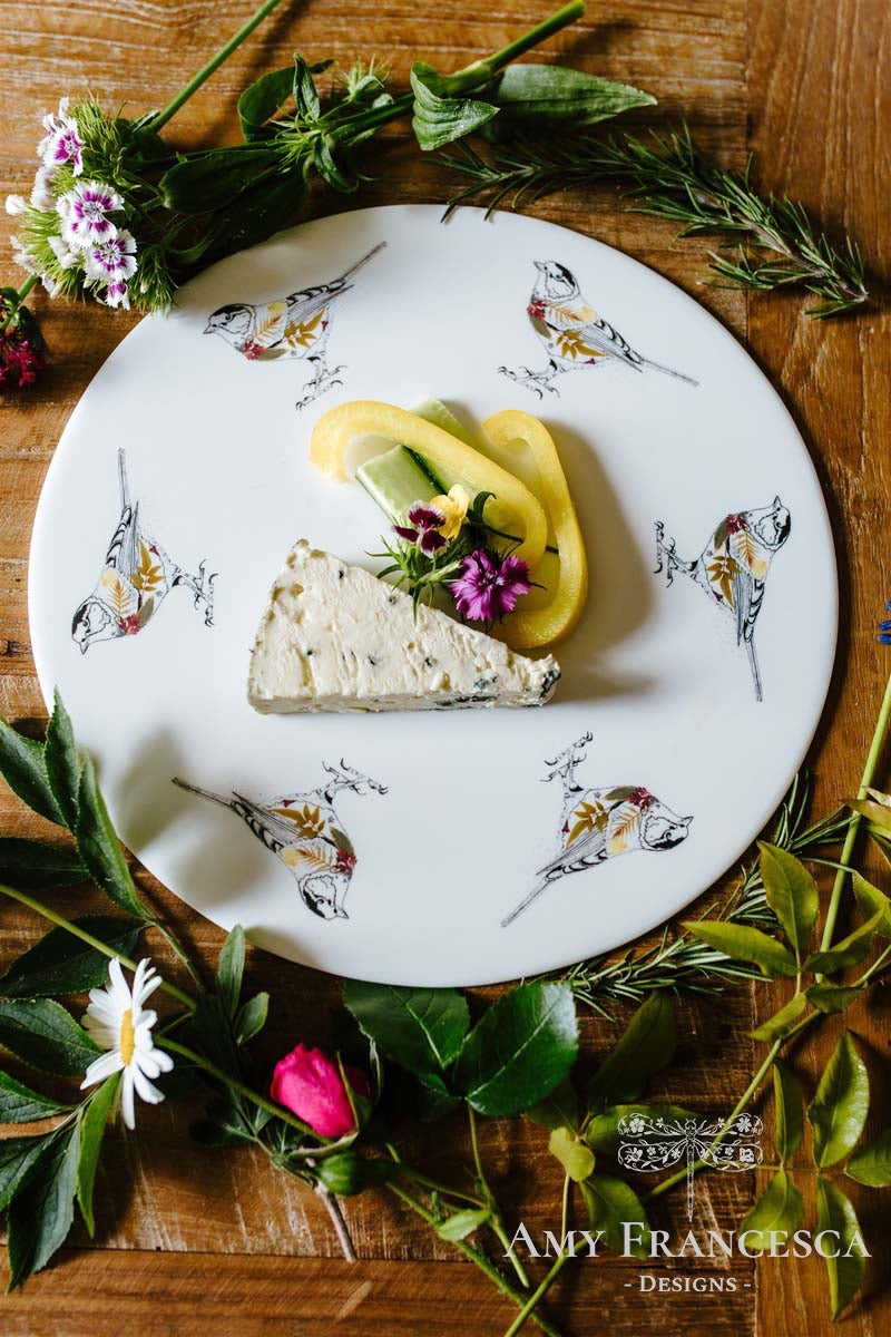 Serving platter featuring bird illustrations, shown with a slice of cheese and surrounding herbs and flowers.