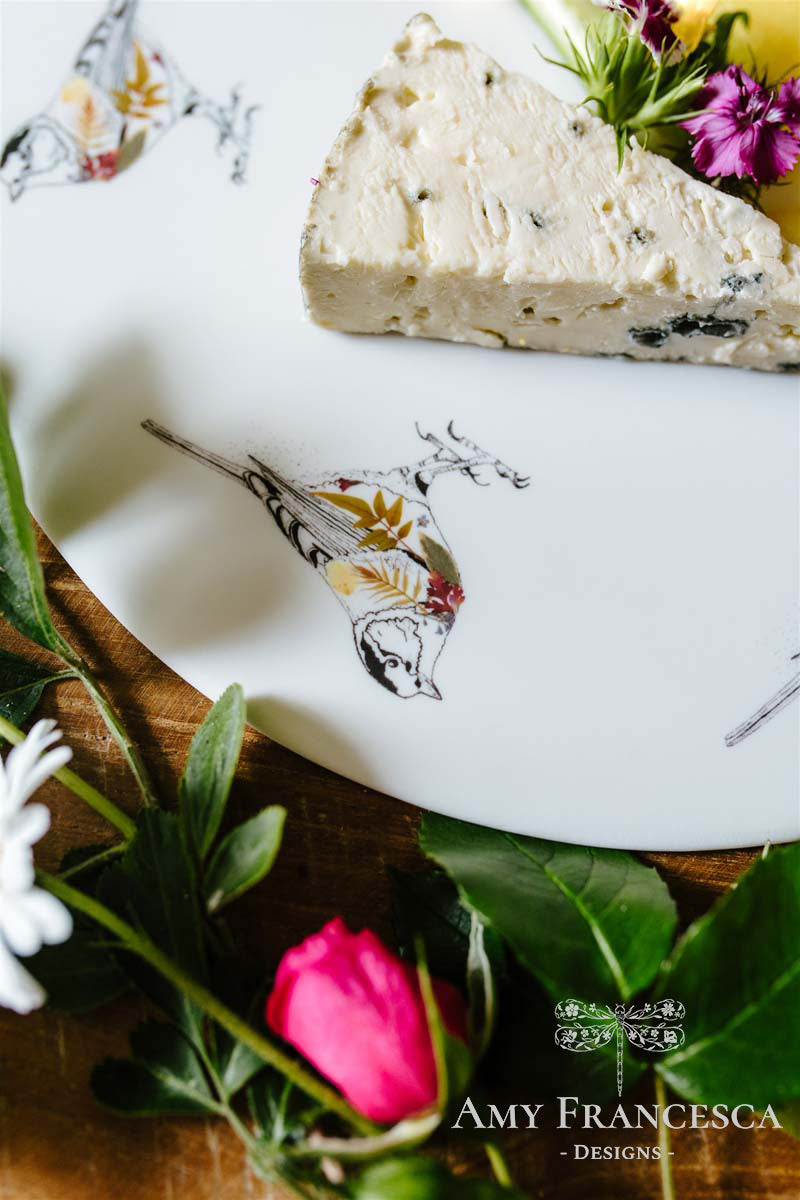 Decorative plate featuring bird illustrations, shown with a wedge of cheese and flowers.