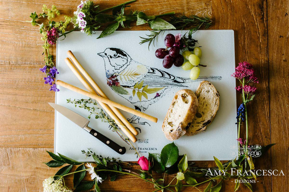 Chopping board featuring a bird illustration, shown with breadsticks, grapes, sliced bread and surrounding flowers.