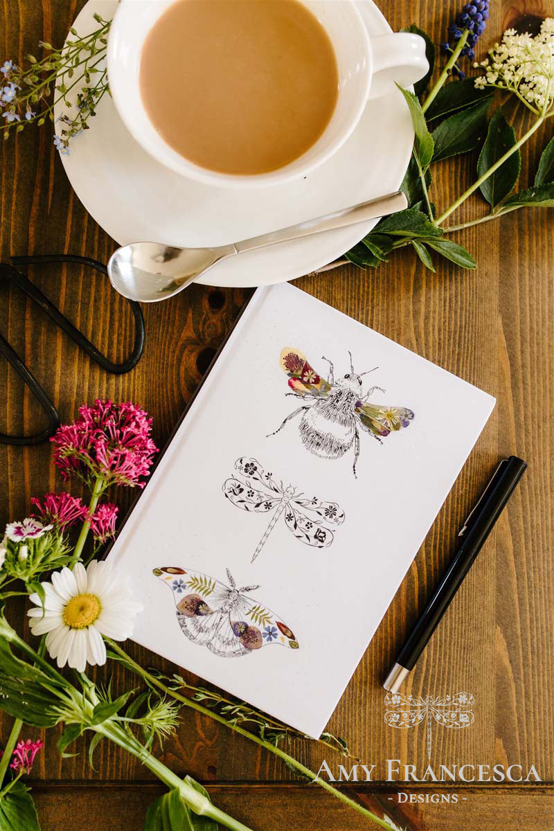 Notebook featuring a bee, a dragonfly and a moth illustration on a white cover, shown with a cup, glasses, a pen and flowers.