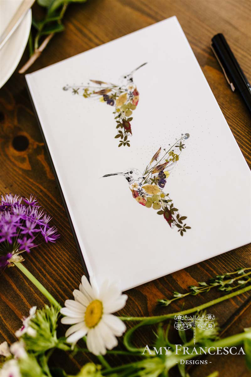 Notebook featuring two hummingbird illustrations on a white cover, shown with surrounding flowers.