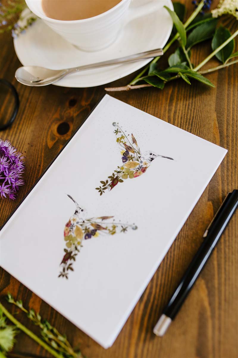 Notebook with two hummingbird illustrations on the cover, placed on a table with a cup and flowers.