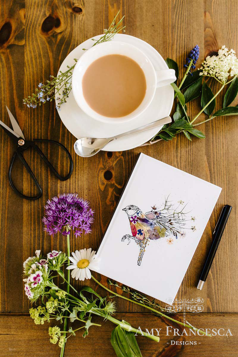 Notebook featuring a bird illustration on a white cover, shown with a cup, scissors, flowers and greenery.