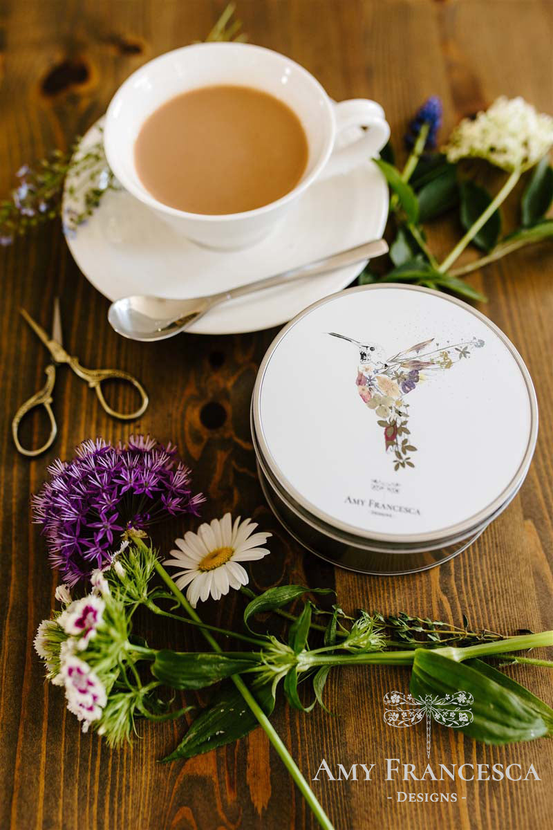 Tin featuring a hummingbird illustration on the lid, shown with flowers, scissors and a cup and saucer.