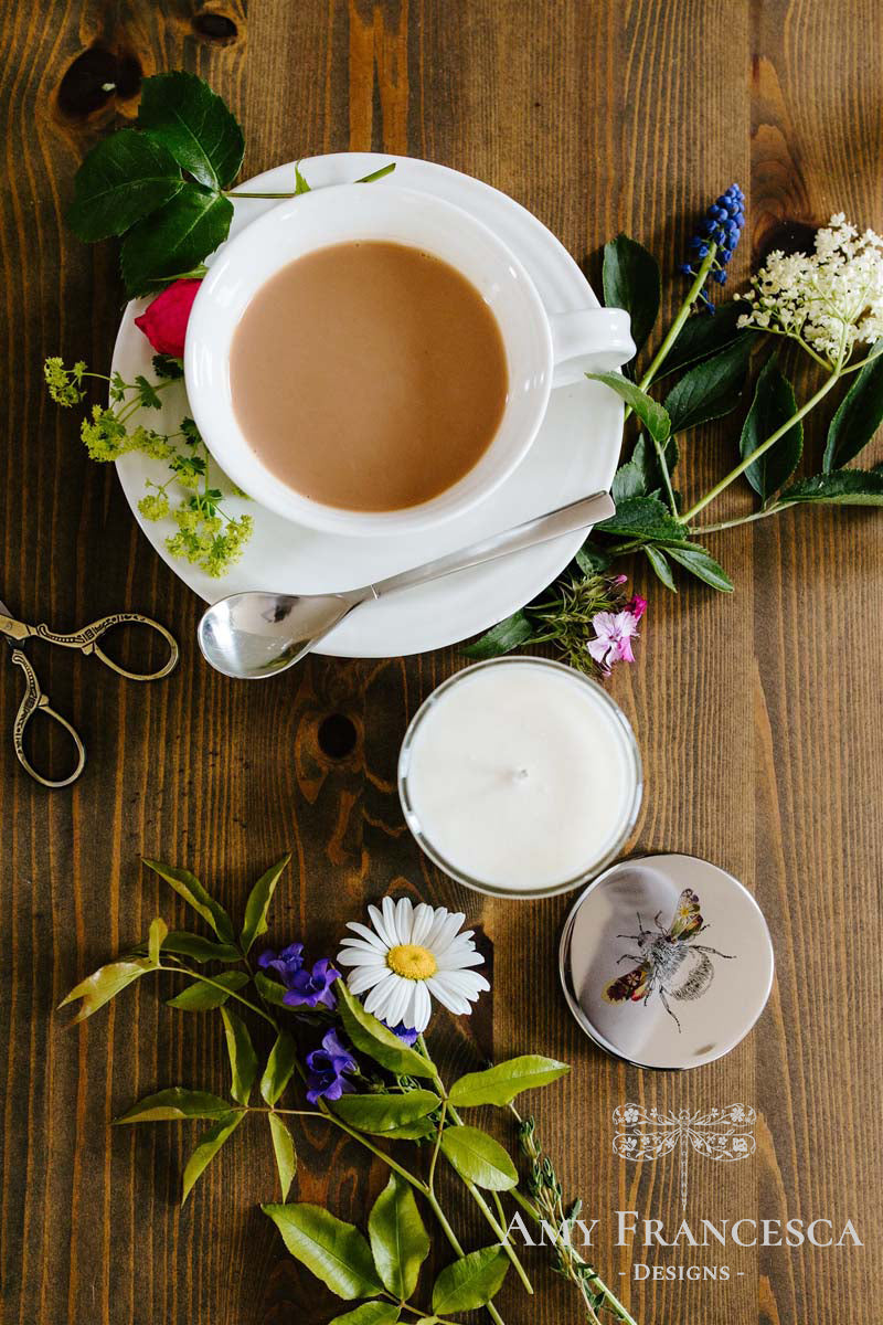 Candle with a bee illustration on the lid, shown with a cup, saucer and surrounding flowers