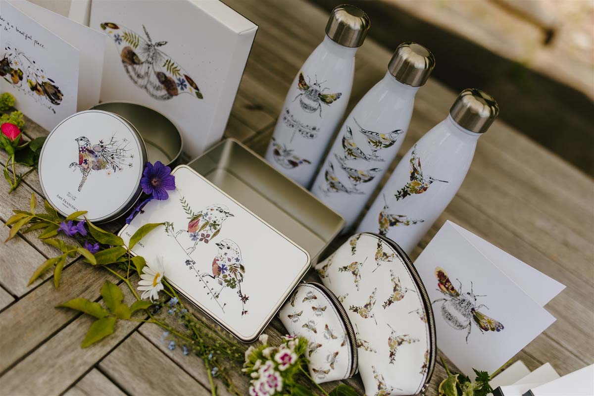 Display of nature‑themed products including illustrated water bottles, pouches, tins and greeting cards arranged on a wooden surface with flowers.