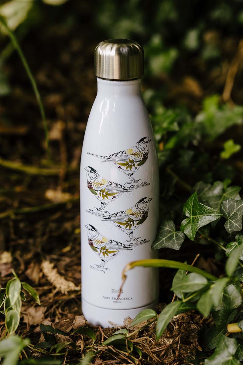 White water bottle with four bird illustrations, photographed among green leaves.