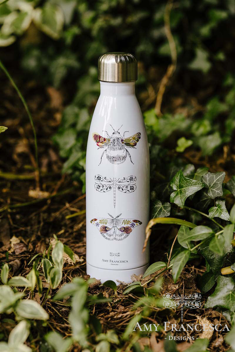 White water bottle featuring bee, dragonfly and moth illustrations, photographed among green leaves.