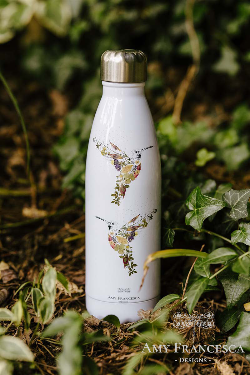 White water bottle featuring hummingbird illustrations, photographed among green leaves.